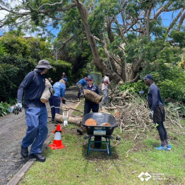 NAB Foundation Recovery Crew volunteers working alongside Disaster Relief Australia clearing debris and green waste from a residential property. They are seen working together, using tools to remove fallen tree branches after a storm event.